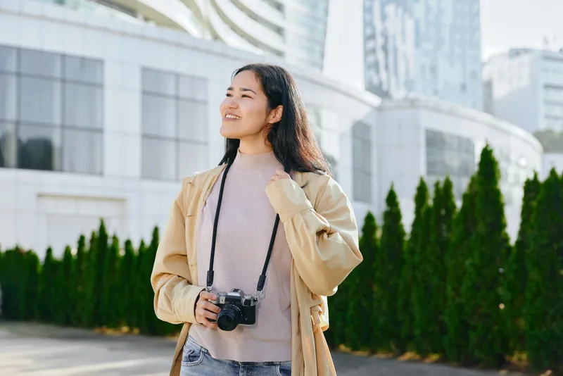 Woman with camera in outdoor urban setting, smiling as she walks past modern buildings, dressed in beige jacket and sweater, capturing moments in daylight