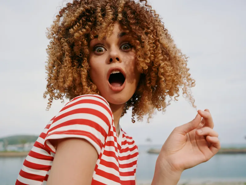 Vibrant portrait of a young woman with curly hair wearing a red striped shirt, eyes wide, mouth open in astonishment, against a seaside backdrop.