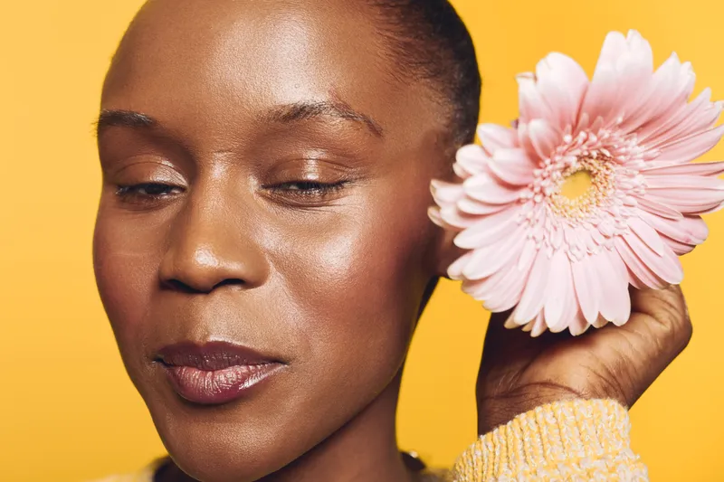 Close-up of a smiling dark skinned African woman holding a pink flower near her face on yellow background. Concept of natural beauty, skincare, and wellness with ethnic female model.