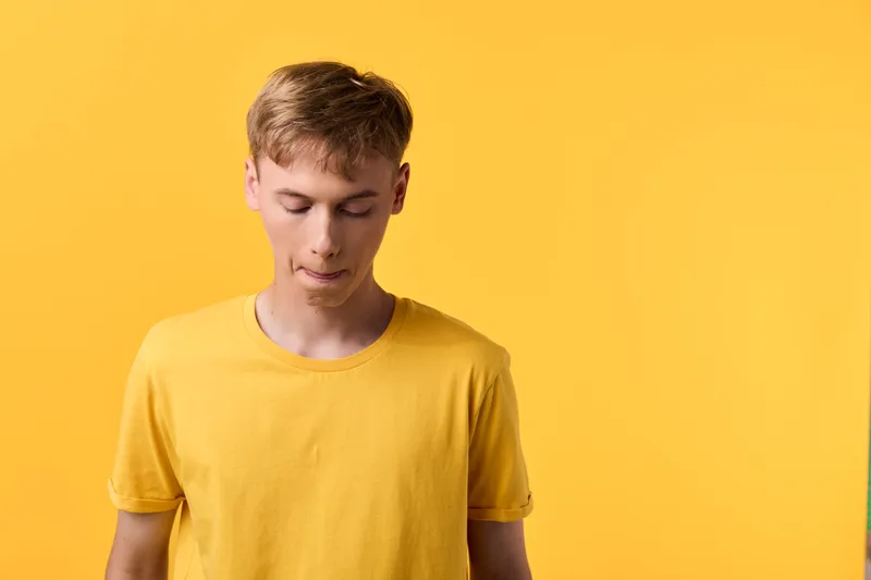 Young man in a yellow shirt stands with a calm expression against a vibrant studio backdrop, showcasing casual style and minimal, contemporary mood