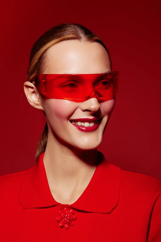 Smiling woman with red sunglasses and red outfit poses against red background. Fashion portrait with vibrant colors and modern style. Concept of confidence and boldness.