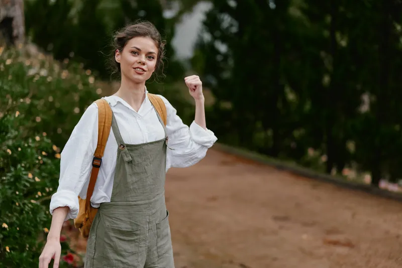 Happy young woman in casual outfit with backpack expressing joy in nature, showcasing vibrant green background and positive emotion