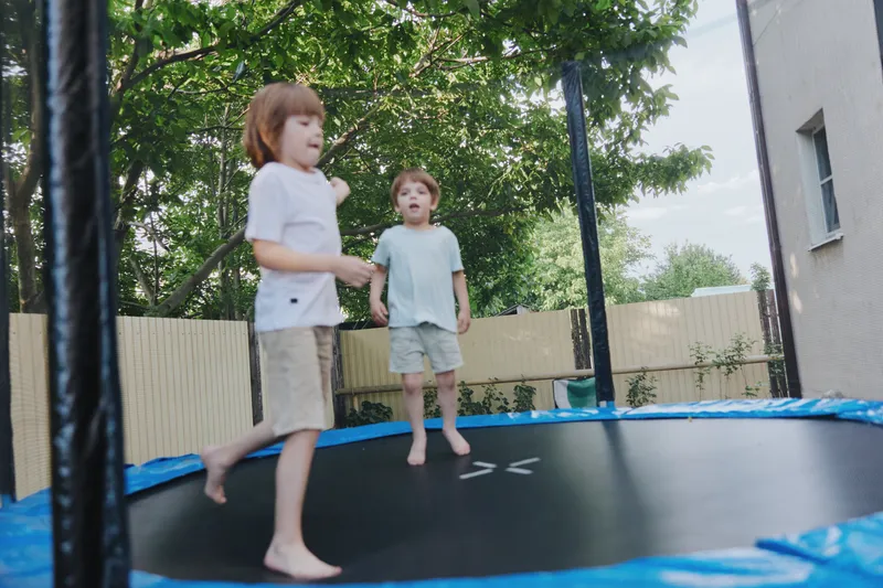 Excited children jumping, surrounded by greenery, on a trampoline in a sunny backyard, showcasing the joy of play and childhood in a vibrant outdoor setting. ###