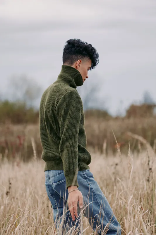 A solitary subject walks through tall meadow grass, dressed in a textured green knit turtleneck and denim jeans, conveying authenticity and grounded veracity.