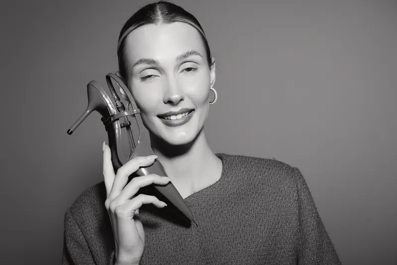 Smiling caucasian woman with light skin holds high heel shoe near face in studio, concept for fashion, style, and footwear.