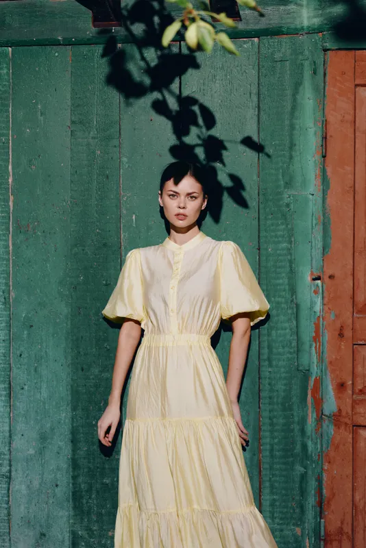 Woman in pale yellow dress stands against a green wall under dappled light, creating a calm fashion portrait with natural ambience and subtle cinematic mood.