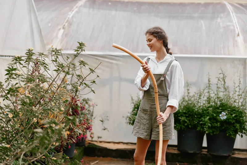 Young woman gardening in a greenhouse, wearing stylish overalls and smiling, surrounded by plants and flowers, capturing the essence of eco friendly living and seasonal growth
