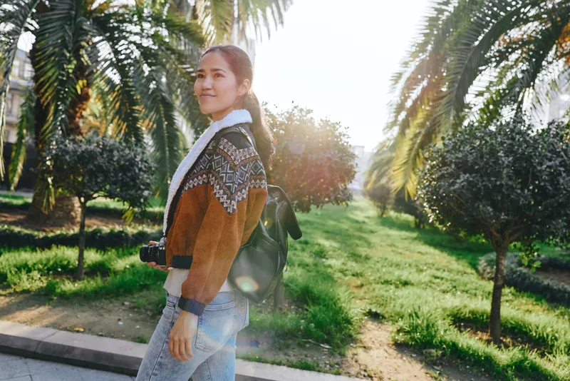 Woman with camera strolling outdoors in a sunny park, wearing a knit sweater and scarf, capturing nature and light during a relaxed afternoon walk, with vibrant greenery and warm glow.