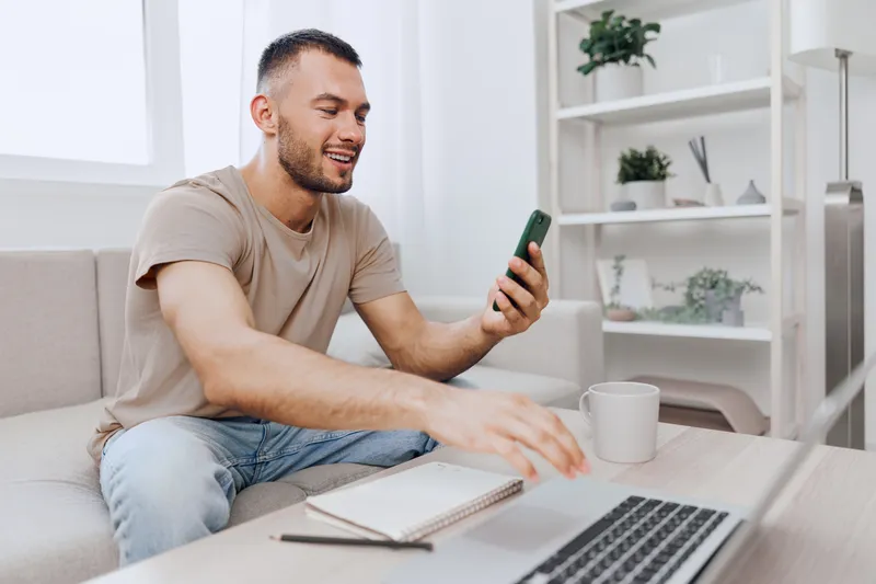 Engaged man smiling while using a smartphone at home, showcasing modern lifestyle, technology, and comfort in a cozy living space with a minimalist design.
