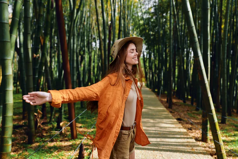 Woman in bamboo forest on a shaded path wearing a straw hat and orange jacket, smiling with arms outstretched, enjoying travel and nature in a serene outdoor portrait and eco tourism scene.