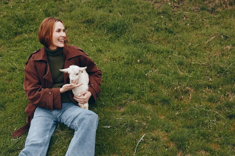 Smiling woman sits on grass holding a kid goat in a park, wearing casual fashion. A warm lifestyle moment that celebrates human animal companionship outdoors, simple rural joy, and a relaxed sense of