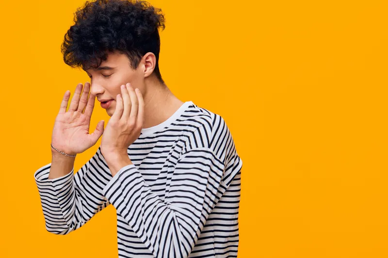 young man in striped shirt whispering with hands near mouth on bright orange background showing secretive gesture and focused facial expression