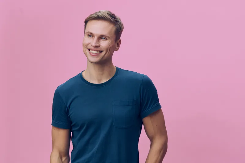 Smiling young Caucasian man in casual blue T-shirt standing against pink background, looking happy and relaxed, showcasing positive emotions and confident demeanor, studio portrait, lifestyle concept,