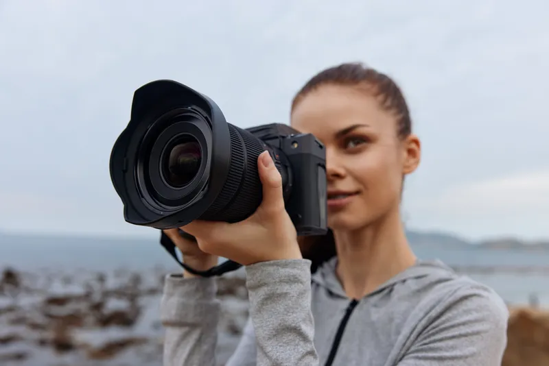 Young woman with long hair capturing nature with a camera, wearing a grey sport outfit, displaying confidence against a coastal background