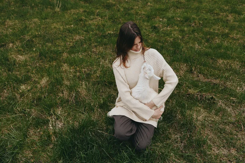 Carefree woman kneels on grass, holding a white goat kid in a cozy sweater, sharing a gentle moment outdoors, showcasing a warm lifestyle scene focused on animal companionship and nature.