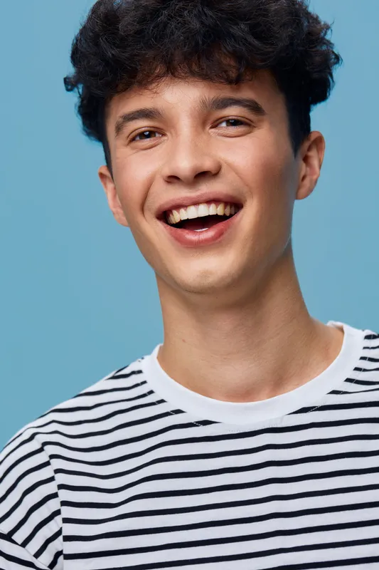 smiling young man wearing black and white striped shirt with curly hair on blue background, joyful expression and casual style in studio lighting