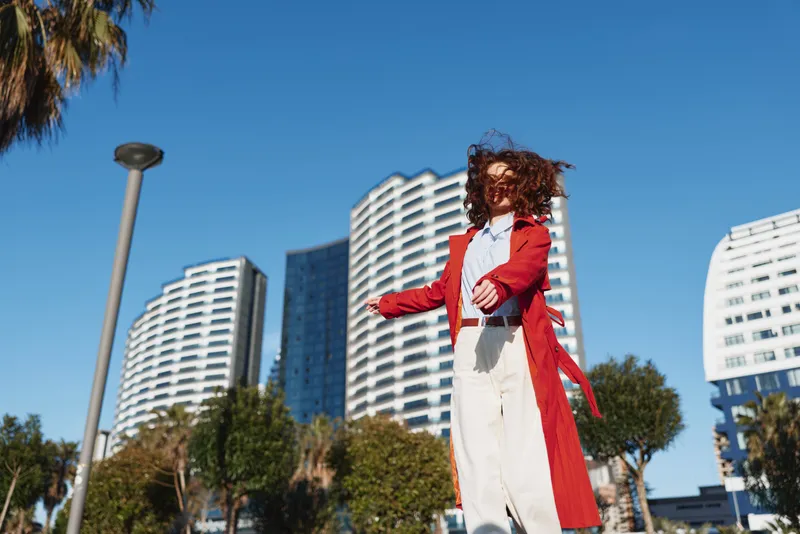 Young woman in a red coat spins joyfully outdoors against modern high-rise buildings and clear blue sky on a sunny day.