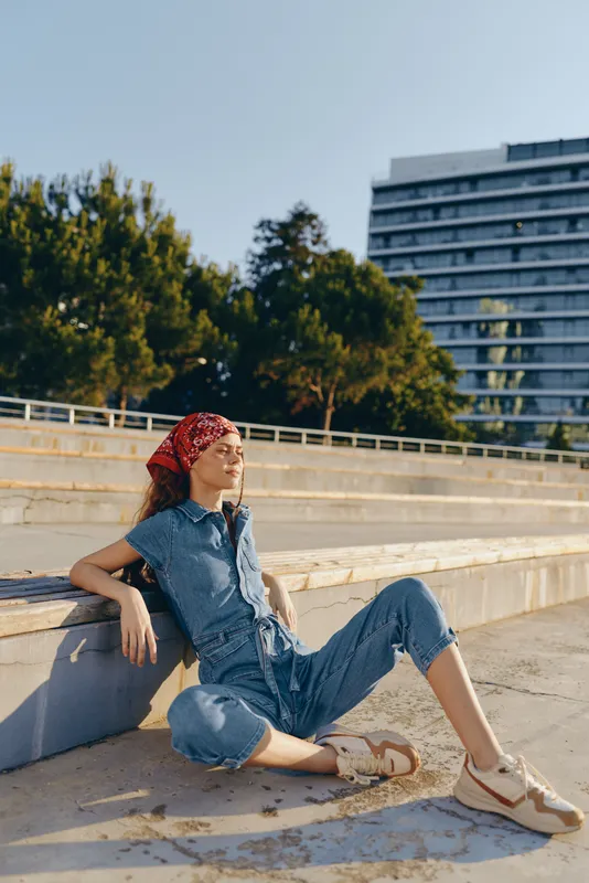Lifestyle woman in modern boho-western denim street style sitting relaxed outdoors during sunny day, film color warm tones, casual urban fashion vibe.