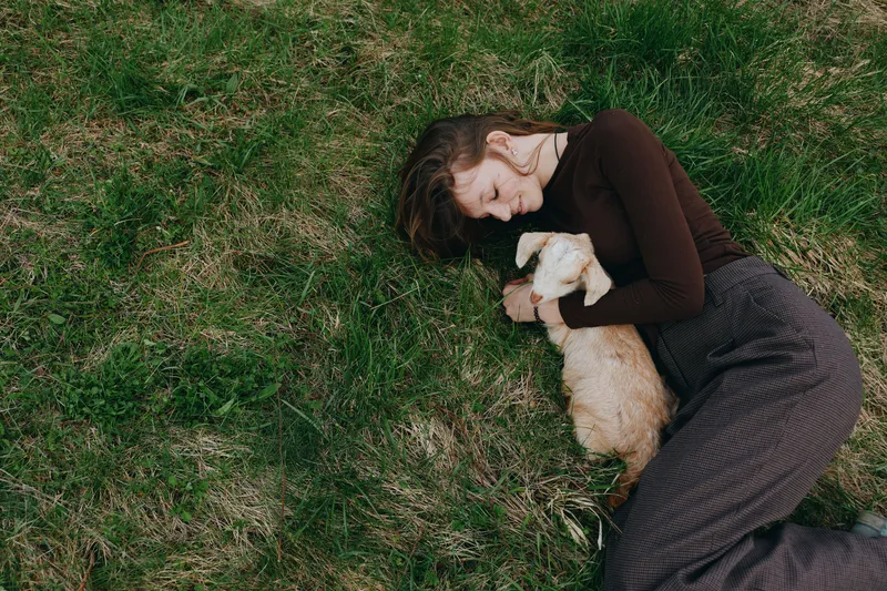 Relaxed woman lying on green grass, embracing a small dog in a sunny outdoor setting, conveying warmth, comfort, and companionship in a casual lifestyle portrait.