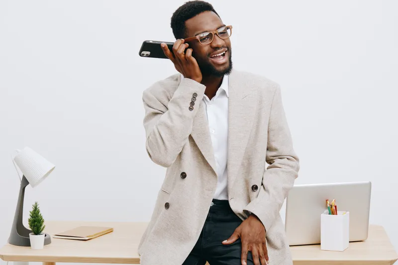 Professional young African American man in smart casual attire talking on mobile phone while standing at office desk, working remotely, on white background. Business communication, technology, office 