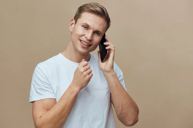 Smiling young man with short hair wearing casual light blue t-shirt talking on mobile phone isolated on plain beige background, studio portrait, people lifestyle concept, happiness, communication, lei