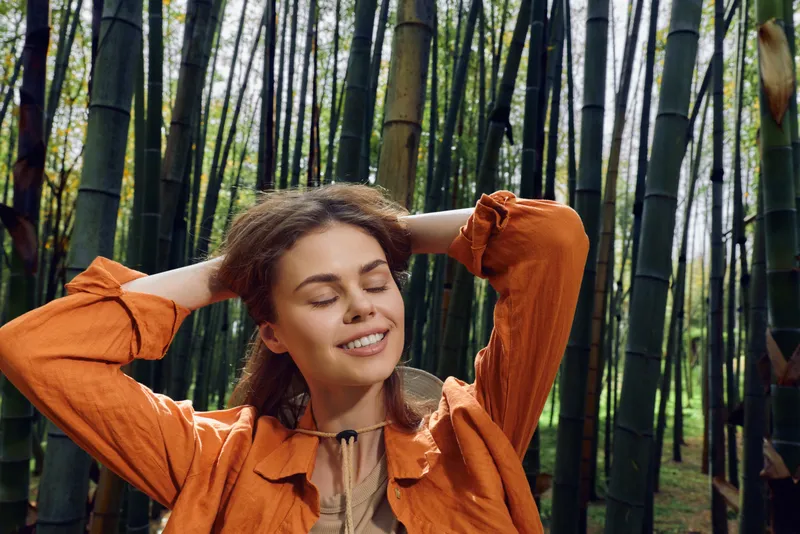 Woman bamboo forest smiling nature portrait relaxation eyesclosed in a tranquil outdoor scene, young adult enjoying sunlight and calm among tall green stalks of bamboo.