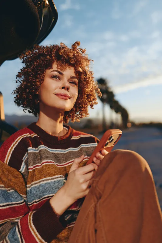 Smiling woman with curly hair holding a smartphone, outdoor lifestyle at sunset, striped sweater, warm golden light, relaxed portrait, modern communication sitting by a path