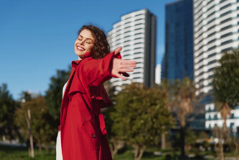 Young woman in a red coat stands outdoors with open arms, smiling in a sunny city park beside tall modern buildings.