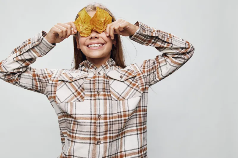 Child smiling and holding two autumn leaves over eyes wearing a plaid shirt on white background. Kid playing with dry yellow leaves and having fun with fall nature indoors.