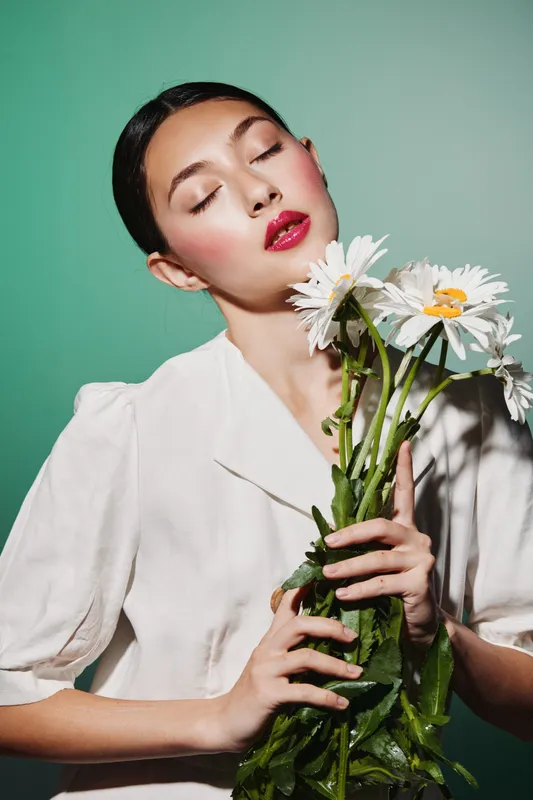 Young woman in white blouse holding daisies against a green background with closed eyes and glowing makeup. Concept of beauty creative fashion and elegant styling.