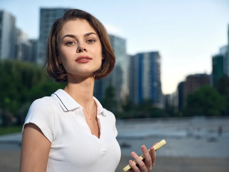 A confident young woman stands outdoors in an urban park, with a city skyline behind her. She gazes at the camera with poise and a calm, approachable energy.
