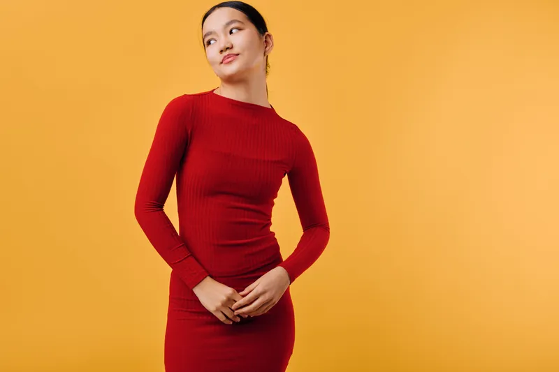 Woman, dress, red, fashion, standing and looking sideways on orange background. Young female model posing with hands clasped for style, beauty and casual outfit concept in studio photo.