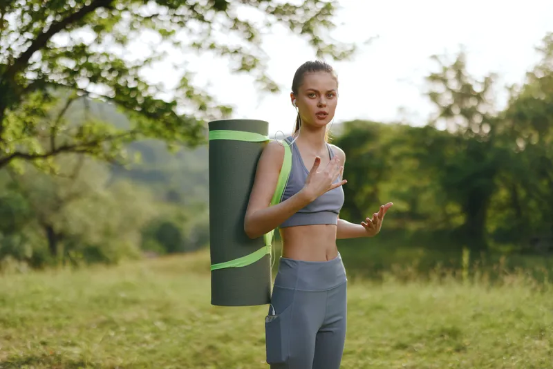 Young athletic woman in a fitness outfit carrying a yoga mat, showcasing determination and empowerment in a lush green park during summer