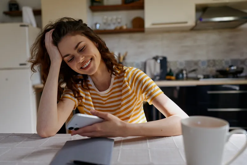 Smiling young woman in a striped yellow shirt relaxes at a bright kitchen, scrolling on a smartphone for a casual home moment
