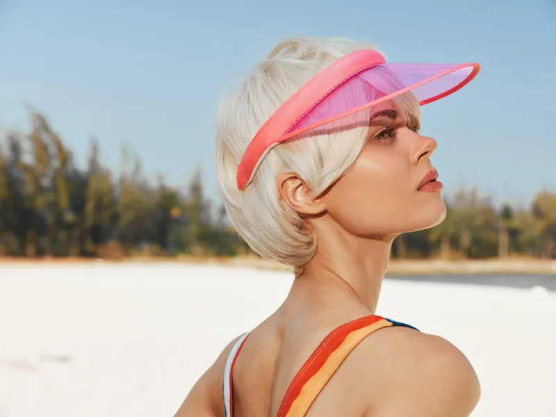 Portrait of a stylish woman with short blonde hair wearing a pink visor at a bright beach, gazing toward the horizon, embodying summer chic and relaxed confidence.