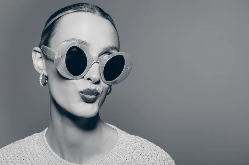Young white woman with pale skin wearing large round sunglasses and hoop earrings poses with pout lips in studio. Concept of fashion, style, and modern accessories.