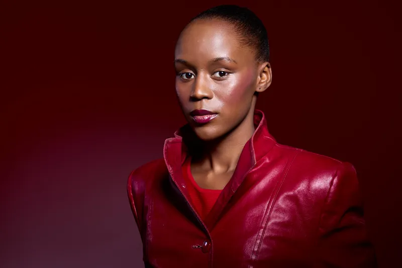 Portrait of elegant black African woman with dark skin wearing shiny red leather jacket and matching top, looking confidently at camera. Studio lighting emphasizes smooth skin and stylish fashion.