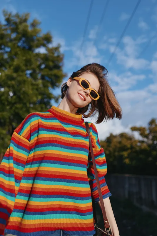 Young woman in rainbow sweater and yellow sunglasses smiling outdoors under blue sky. Early twenties model in casual outfit with shoulder bag, windblown hair and cheerful lifestyle portrait vibe.