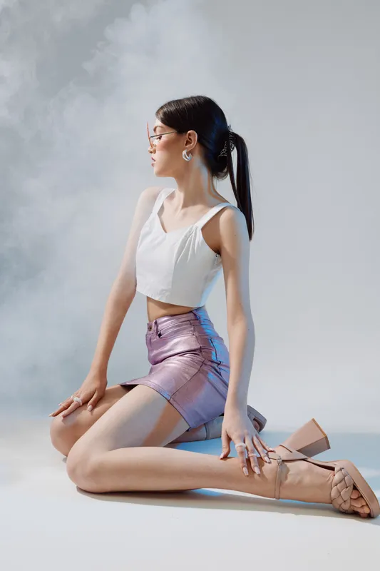 Young woman in a white crop top and shiny skirt poses kneeling on the floor. She wears glasses and heels with a sleek ponytail in a studio setting. Concept of beauty fashion style.