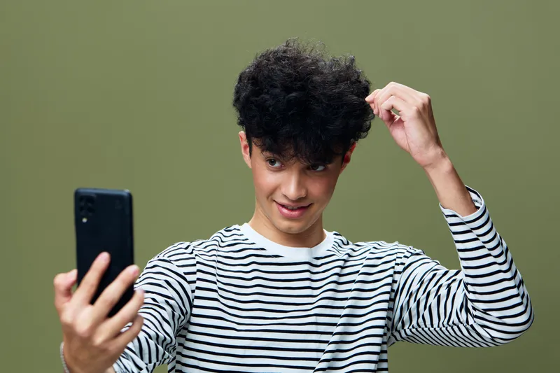 Young man taking selfie with smartphone, wearing striped shirt, smiling and adjusting curly hair in front of olive green background, casual style concept.