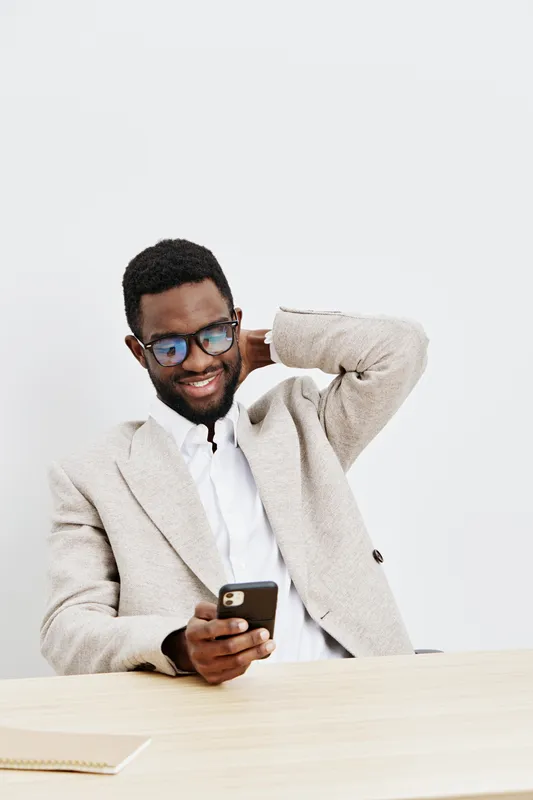 Smiling happy young man with afro hairstyle wearing glasses and beige blazer sitting at desk using smartphone, isolated on plain white background. Casual, cheerful, adult, elearning, technology, lifes