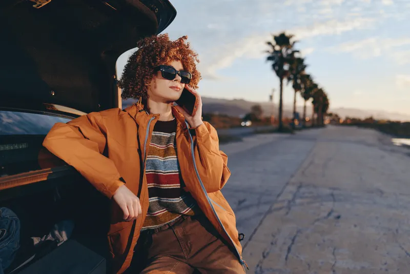 Woman with curly hair and sunglasses, standing by a car on a street, wearing a brown jacket and striped sweater, outdoor urban lifestyle at sunset, warm light, palm view, relaxed mood