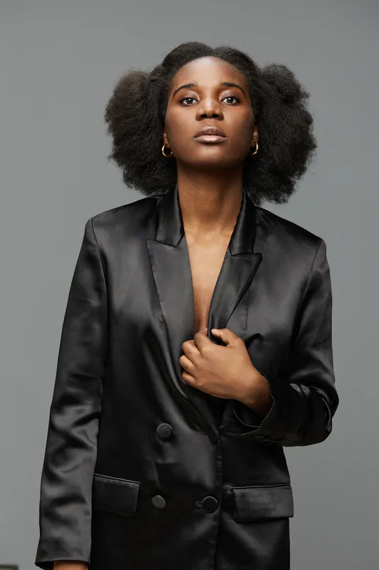 Stylish African American woman in a black satin blazer, exhibiting confidence against a minimalistic gray backdrop Key elements include fashion, bold expression, and modern aesthetics