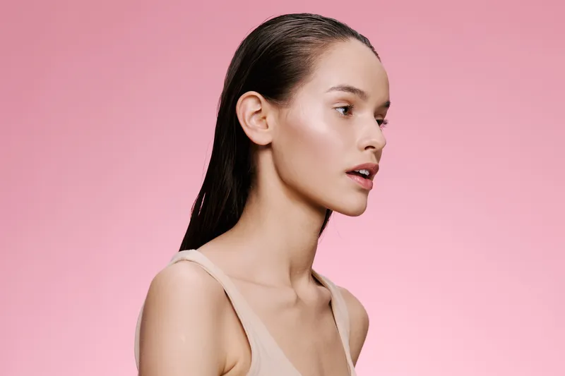 Profile portrait of a beautiful young woman with wet hair against a soft pink background, showcasing natural beauty and minimal makeup