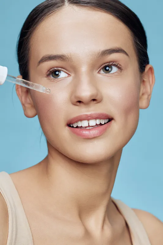 Young woman with a natural glow applying serum on her face, showcasing healthy skincare routine, vibrant blue background, emphasizing beauty and freshness