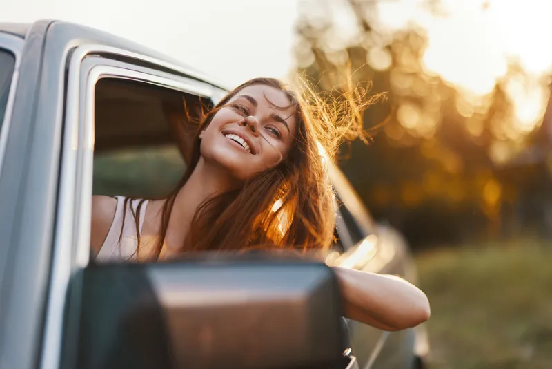 Happy young woman enjoying a road trip with wind in her hair, smiling from a car window during sunset, conveying freedom and adventure.