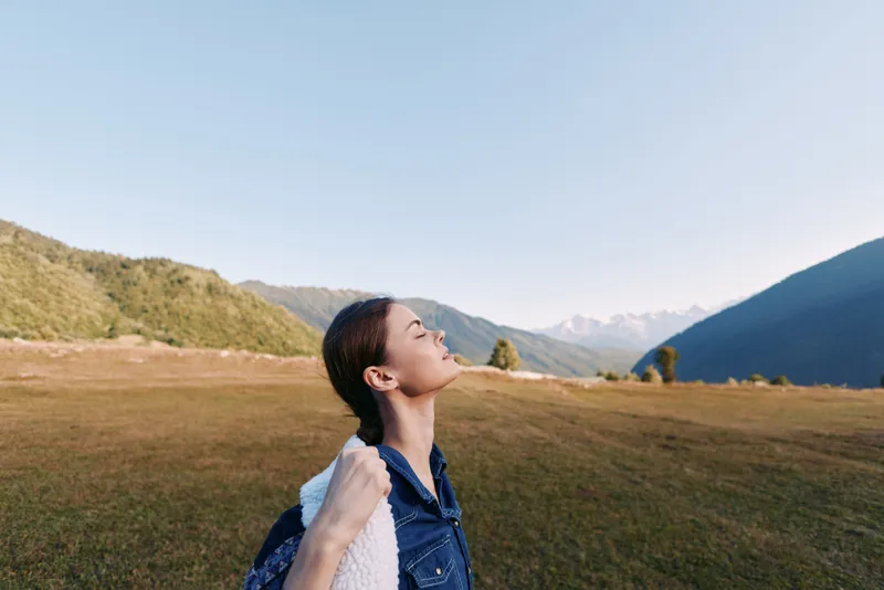 Woman with backpack in mountain meadow enjoying fresh air and nature, hiking and travel portrait with closed eyes, peaceful scenic landscape and relaxed outdoor adventure.