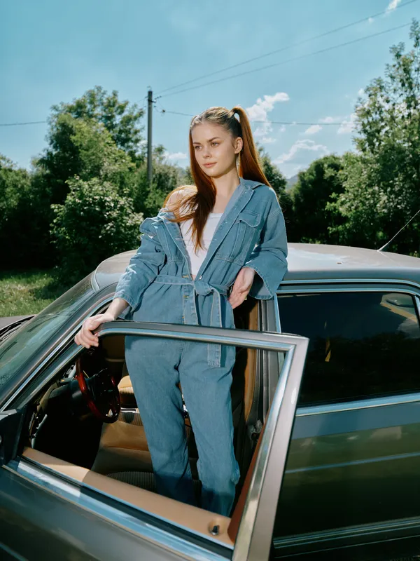 Young woman in denim jumpsuit standing by vintage car in nature, showcasing confidence and style against blue sky and greenery.