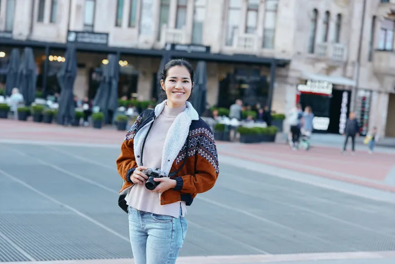 Smiling woman with camera in outdoor city square, casual fashion and urban scenery, candid daytime moment amid daylight, street, architecture and plaza atmosphere