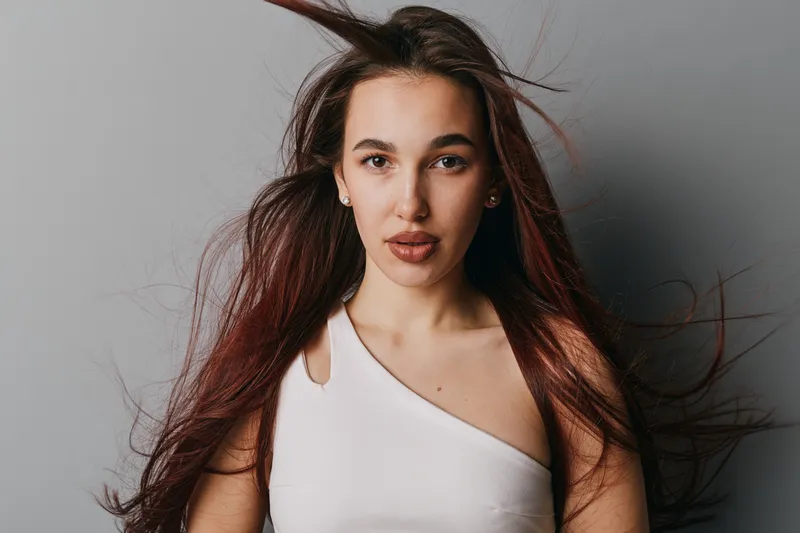 Young woman with long brown hair and natural makeup posing in white sleeveless top against gray background with hair flowing, confident expression and direct eye contact with camera.